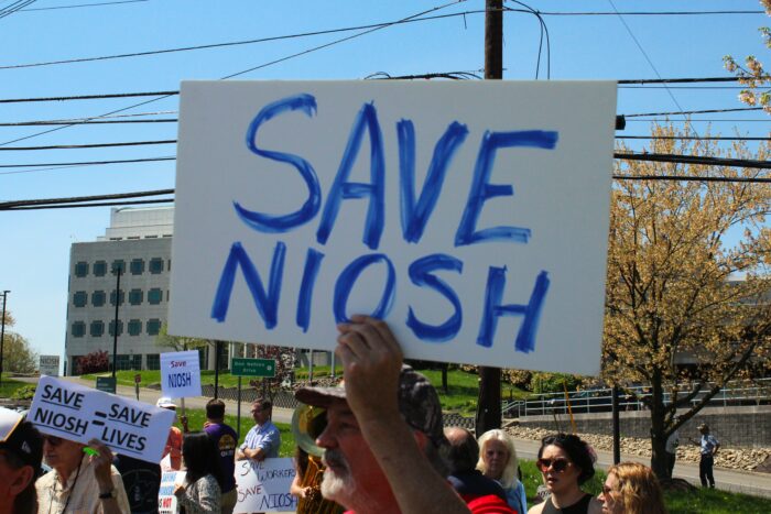 A white sign with blue text reads "Save Niosh" held up by a man with a white goatee over a small crowd
