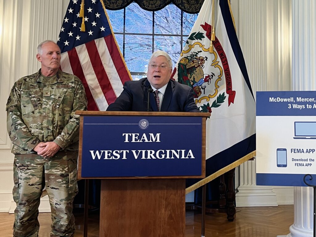A man in a suit and tie stands behind a podium and speaks into a microphone. Behind him stand the American and West Virginia flags, and a military serviceman in uniform. On the other side, a poster outlining ways to access federal disaster aid is on display.