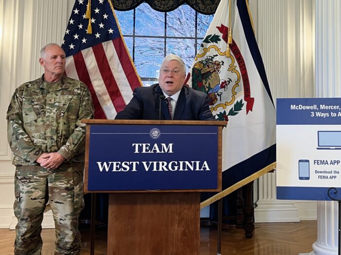 A man in a suit and tie stands behind a podium and speaks into a microphone. Behind him stand the American and West Virginia flags, and a military serviceman in uniform. On the other side, a poster outlining ways to access federal disaster aid is on display.