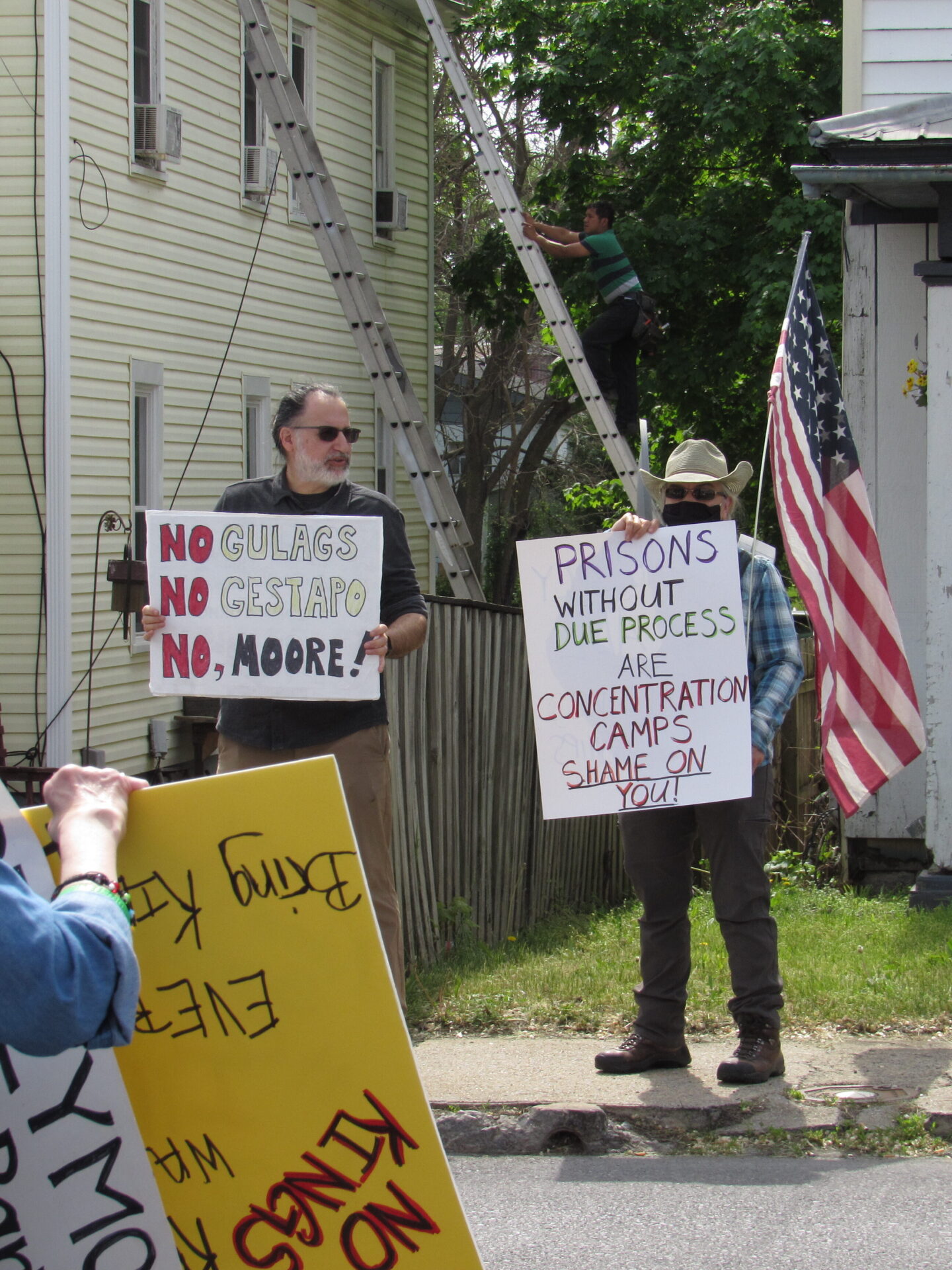 Two people along a sidewalk stand with an American flag and picket signs opposing the detainment of migrants to the U.S. in a notorious El Salvador prison.