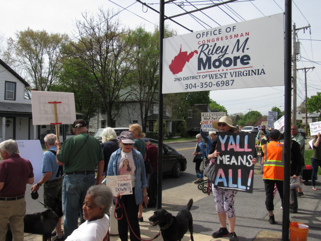 People holding picket signs line a sidewalk beside a sign that reads "Office of Congressman Riley Moore."
