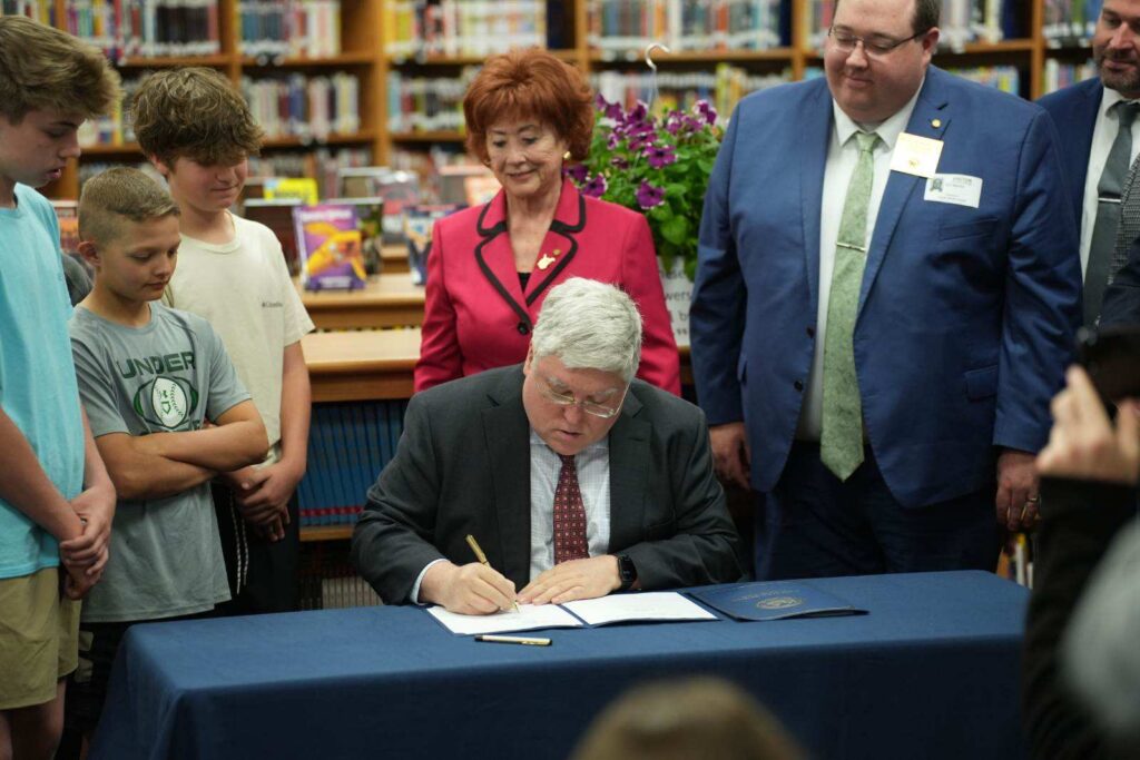 In a school library, a man in a suit and tie sits at a table and signs a piece of paper. On his left stand middle school-aged children. On his right stand other adults dressed in formal attire. Those around him are looking him down, watching him sign.
