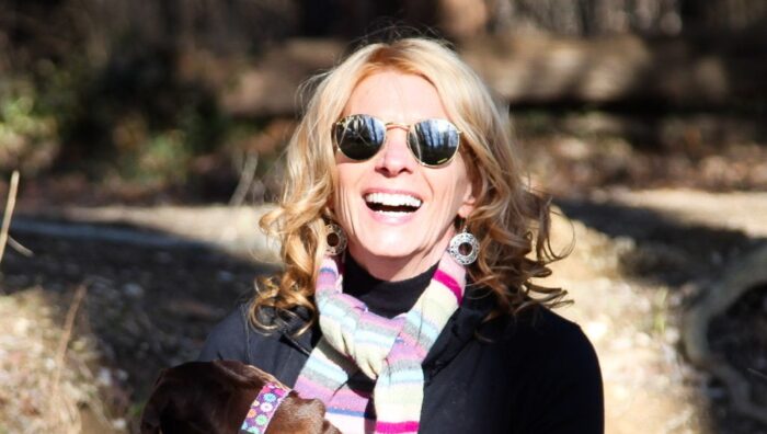 A smiling blond woman in sunglasses sits on a rock with a dog in her lap