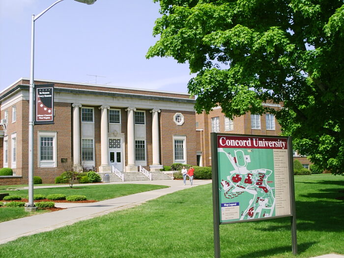 A neoclassical building in the background of the image is the focus of the image. It is build with red brick and has four ionic columns highlighting its entryway. In the foreground the canopy of a large tree dominates the right of frame, directly above a map of the Concord University campus. Two people can be seen walking on a path in the midground in front of hthe building entrance.