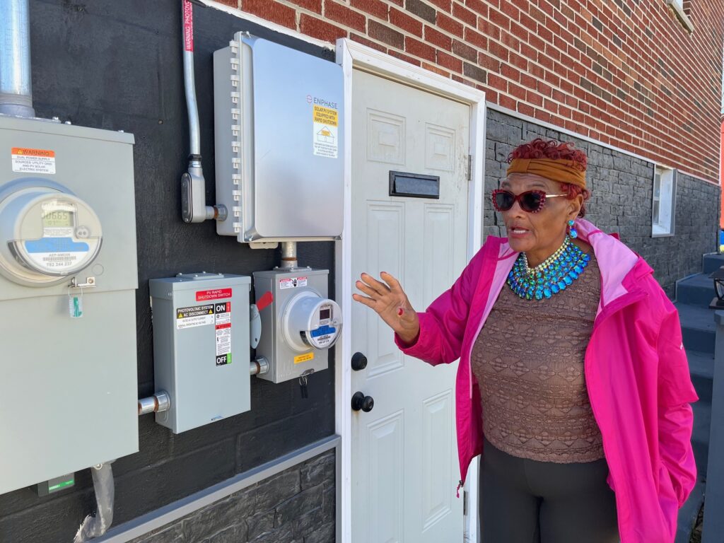 A woman wearing a hat and sunglasses and a pink jacket gestures toward the meters on the side of her red brick house.