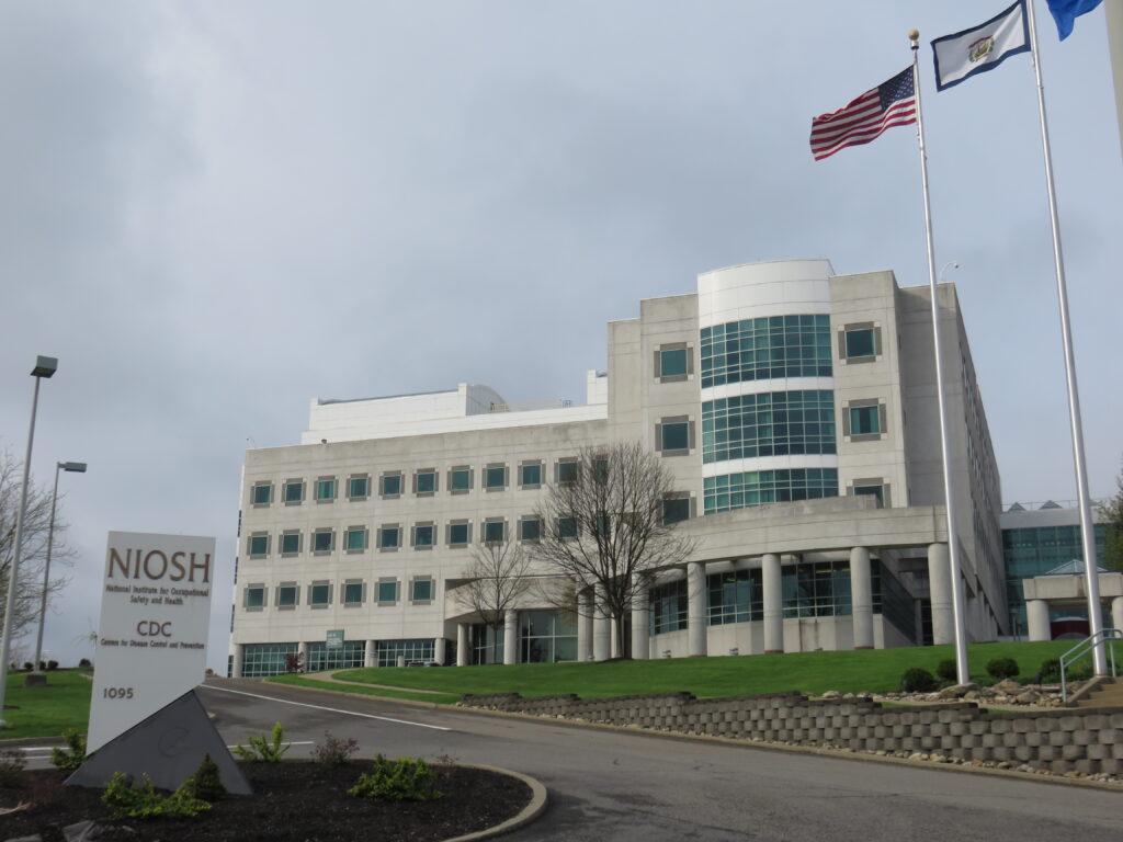 A mulit-story concrete building with rows of circular windows is poised above a driveway and small green lawn. The building is flanked by flags to its right, including the American flag and the flag of the state of West Virginia. A sign on the right of the frame reads "NIOSH National Institute for Occupational Safety CDC Centers for Disease Control 1095"