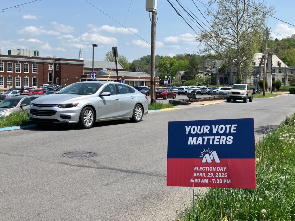 A sign on the side of the road reads "Your Vote Matters" in white on a blue section above a red field with more information about the local Morgantown election April 29. Behind a sign cars are parked next to a high school.