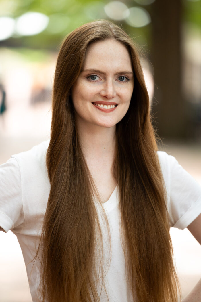 Portrait photo of a white woman with straight, long brown hair wearing a white shirt who is standing outside.