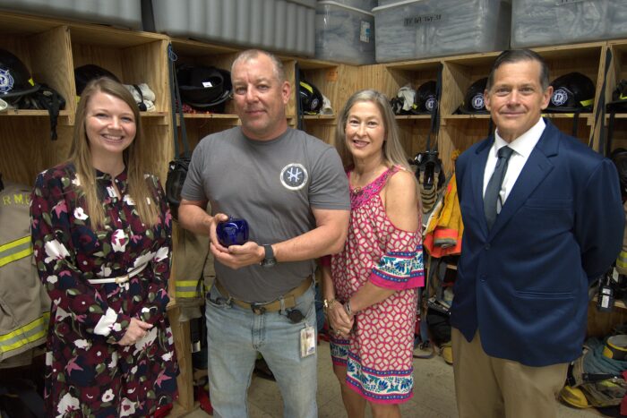 Four adults, two women and two men, pose for a photo. They stand in front of firefighting gear.