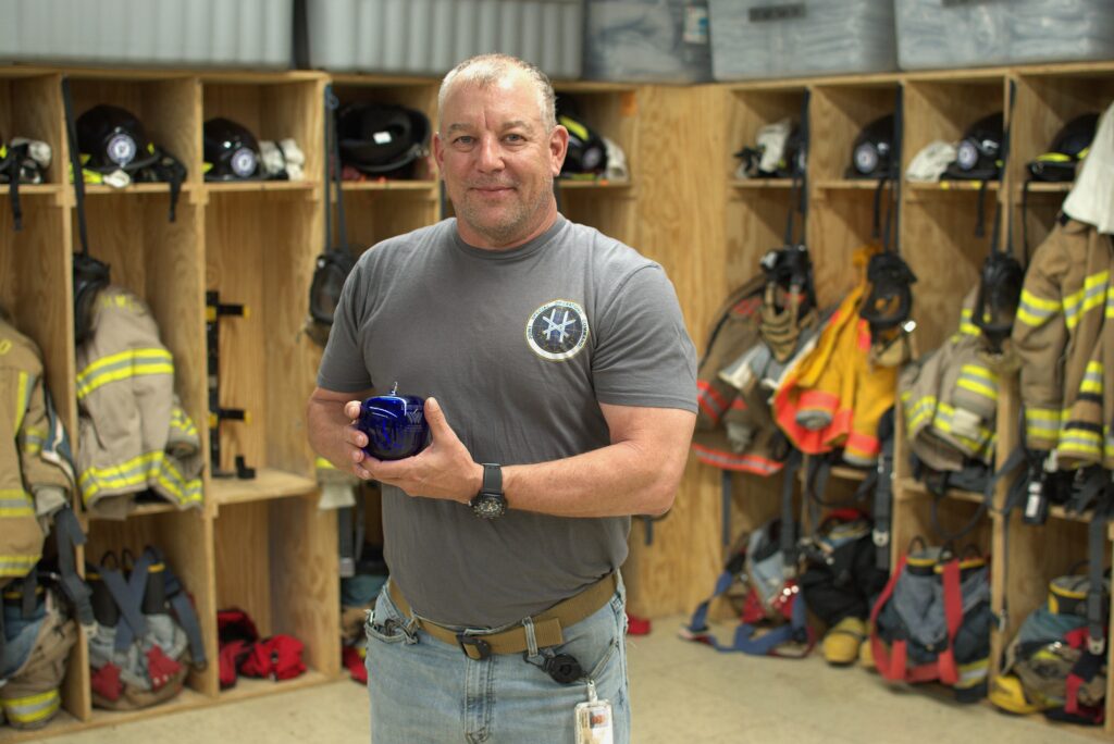 A man stands in front of firefighting outfits and equipment. He holds a blue apple paperweight made of glass.