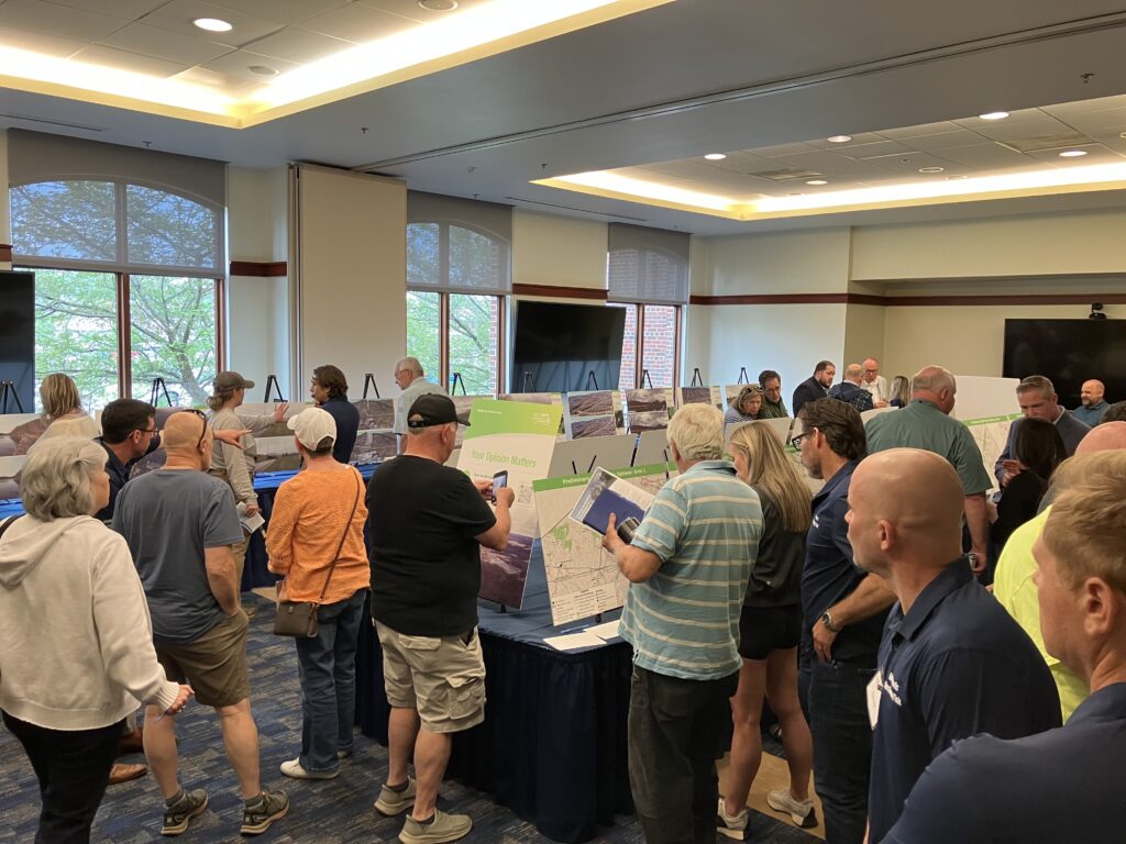 A large group of people stand in a meeting room below fluorescent lights, looking at poster boards arrayed on tables.