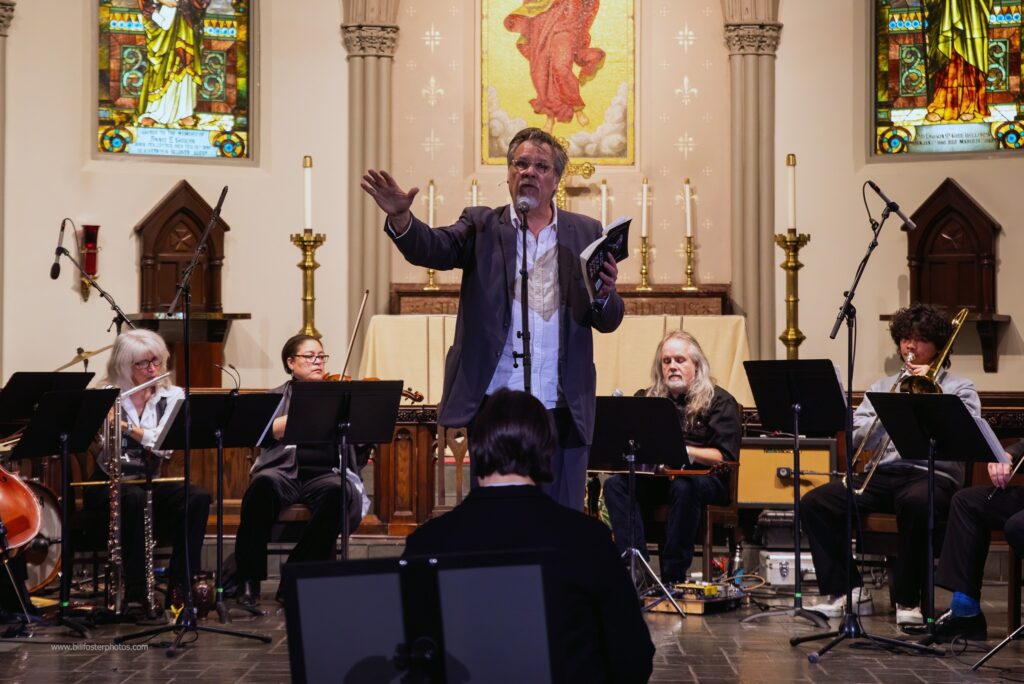 A white man standing in a church with a book in his left hand and speaking into a microphone. There are four musicians playing instruments behind him on a stage and one person sitting in front of him.