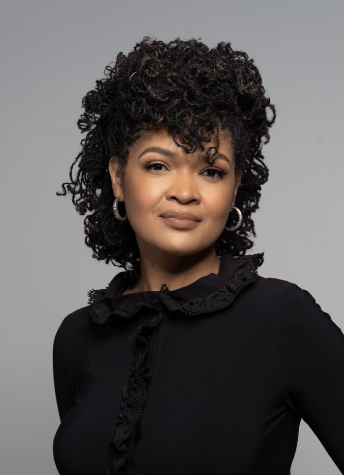 A Black woman posing for a photo. She is wearing a black shirt and standing in front of a gray background.