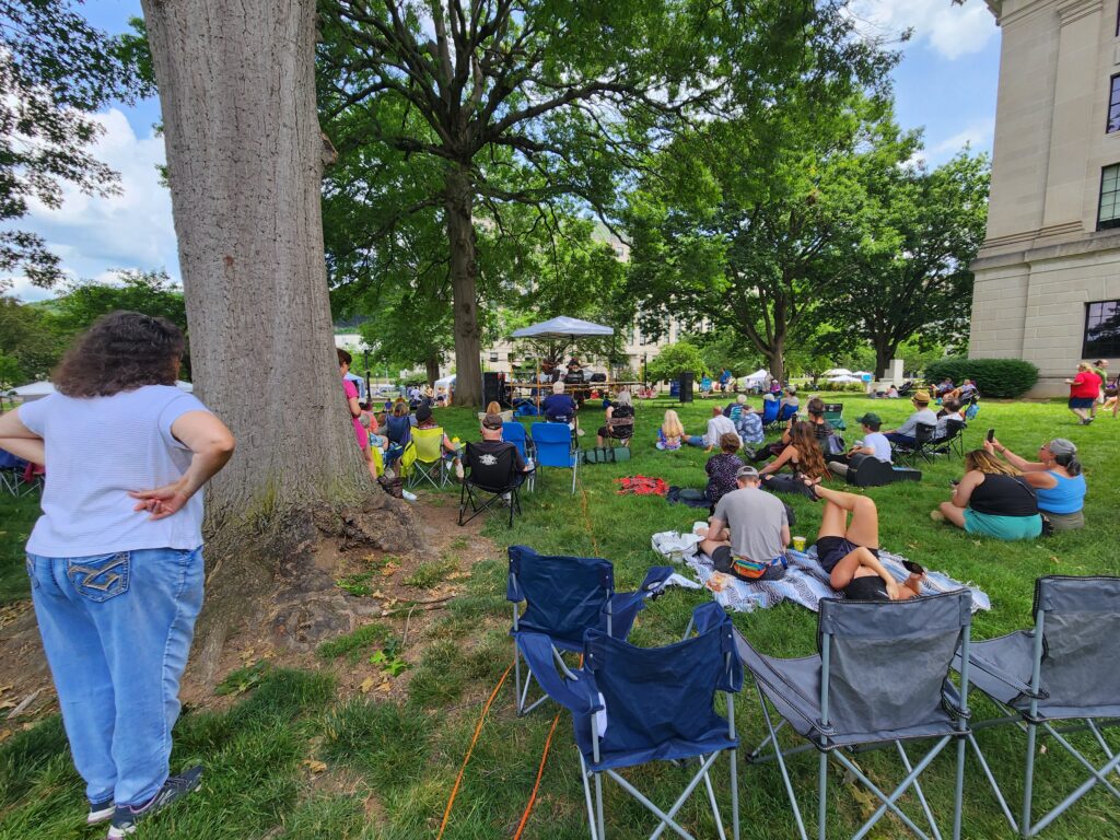 People standing and sitting outside under a tree listening to live music.