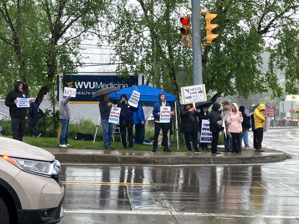 A group stands at the corner of a busy intersection holding signs that read "Tell Capito Restore Niosh" and "WV Needs Good Jobs. NIOSH Needs Its Scientists". Demonstrators wear rain jackets and stand in front of a collapsible blue tent awning.