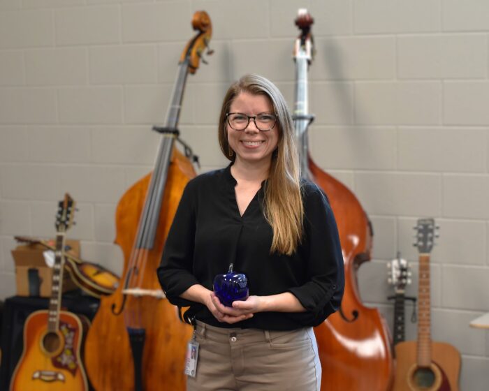 A woman stands in front of cellos, wearing a black dress shirt and slacks. She is smiling and wearing glasses and has long brown hair. She is holding a blue apple Blenko Glass paperweight.