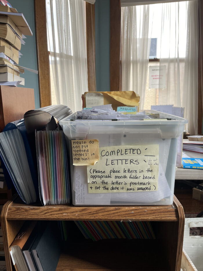 A box of letters on a shelf. The box is in a room stacked with other books and files.