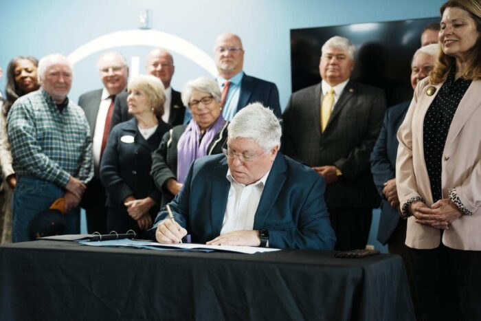 A man wearing a dark blue suit over a white button up shirt with an open collar and no tie sits at a table with a black tablecloth. He grips a pen in his right hand as he signs a paper in a folder. Behind him several people in suits and formal clothes stand arrayed.
