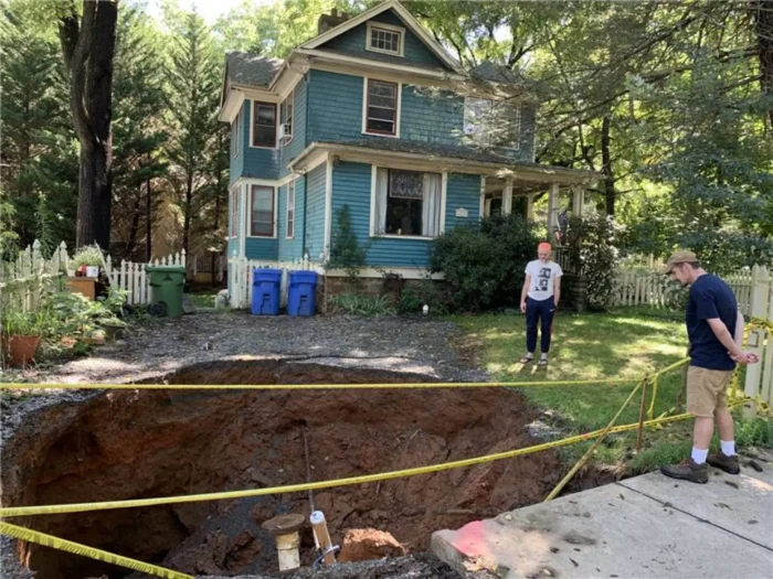 A sinkhole opened up in front of a blue house. Two people are looking into the sinkhole. 
