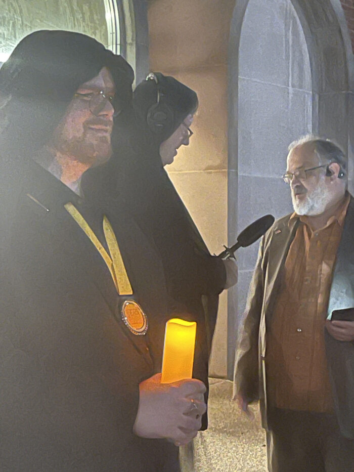 Three men are shown. Two are hooded, and one wears a lanyard with a medal. The other hooded figure is holding a microphone and interviewing an older man with white hair and a white beard.