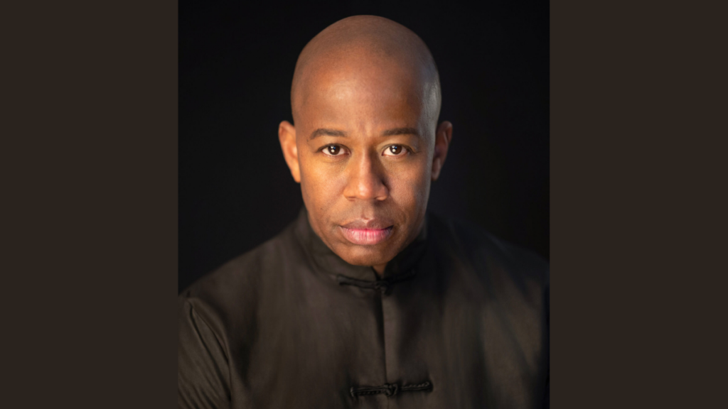 A man, Maestro André Raphel, with no hair stares at the camera wearing a black top.