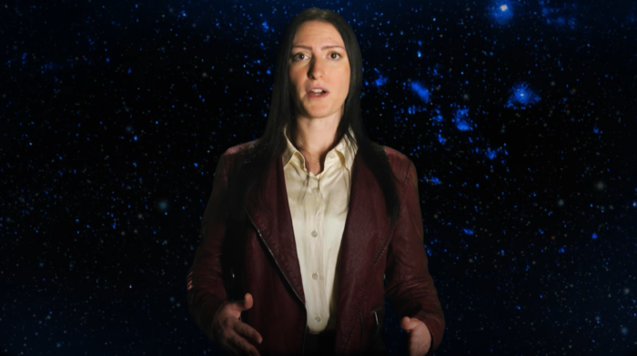 A bust shot of a woman who is speaking. Behind her are stars as we see them from Earth.