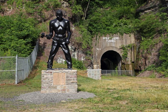A statue of John Henry is shown against a grassy hill and a train tunnel.