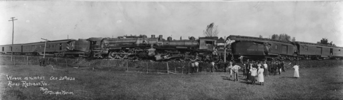 A black and white panoramic photo of a train wreck. Several people are seen standing on the side of the tracks looking up at the wreck.