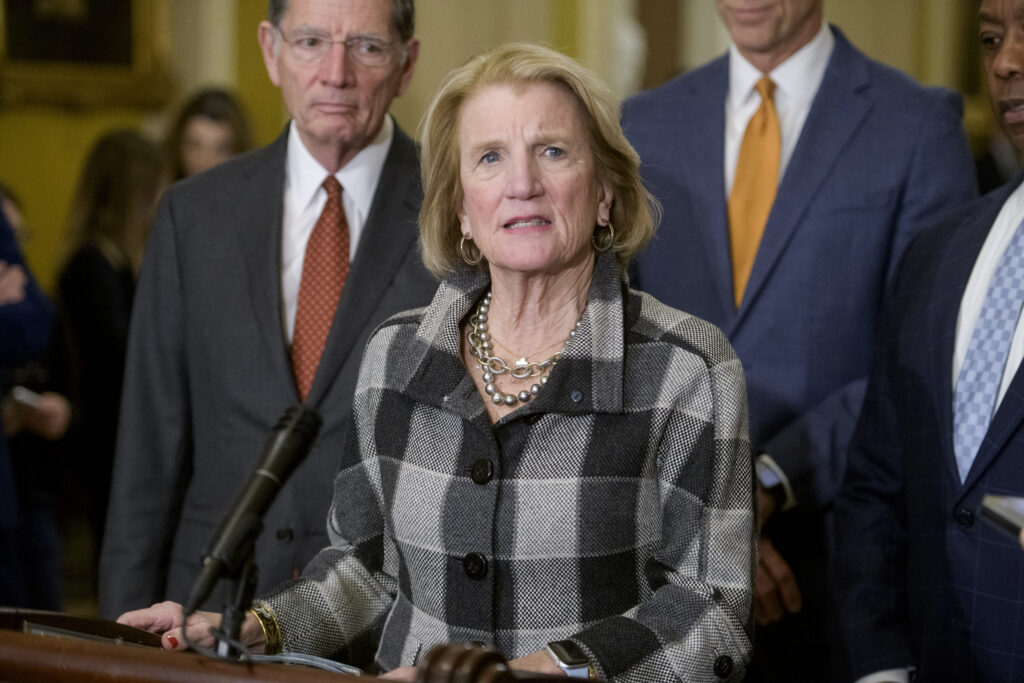 A white woman with blond hair stands a podium. She wears a black and gray checkered button up shirt and hoop earrings. Three men stand behind her.