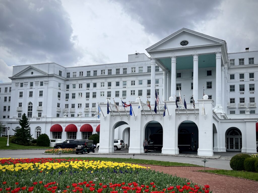 A sprawling white building with a columned entryway sits at the end of a paved loop for cars, with a garden of colorful flowers in front of it.