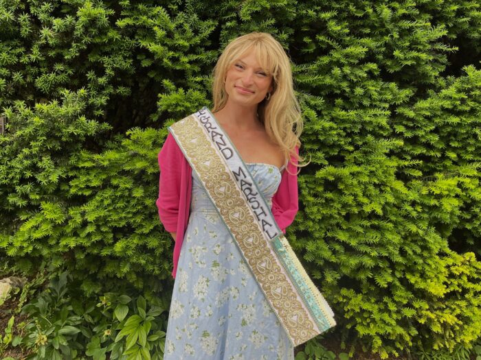 A woman wearing a dress and a sash stands in front of a bush and smiles at the camera.