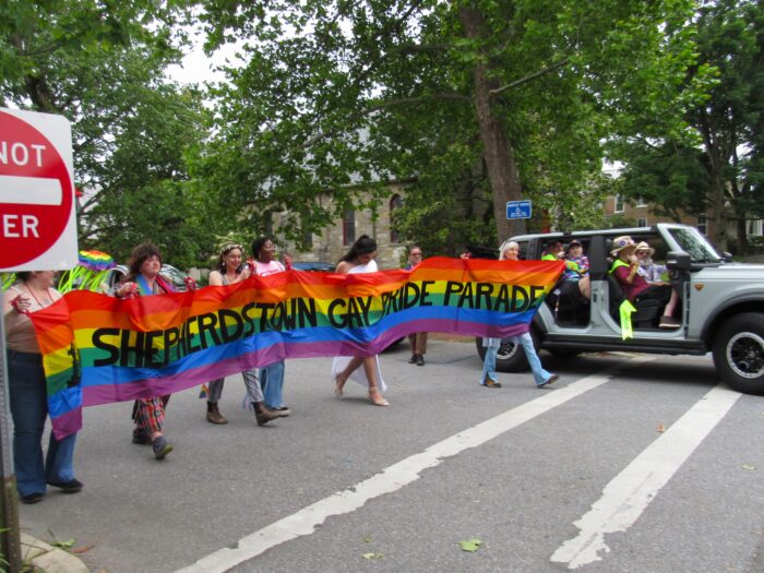 Several people hold rainbow banner, stretching it across the street, and begin to walk. Beside them, a car filled with people wearing sashes starts to drive.