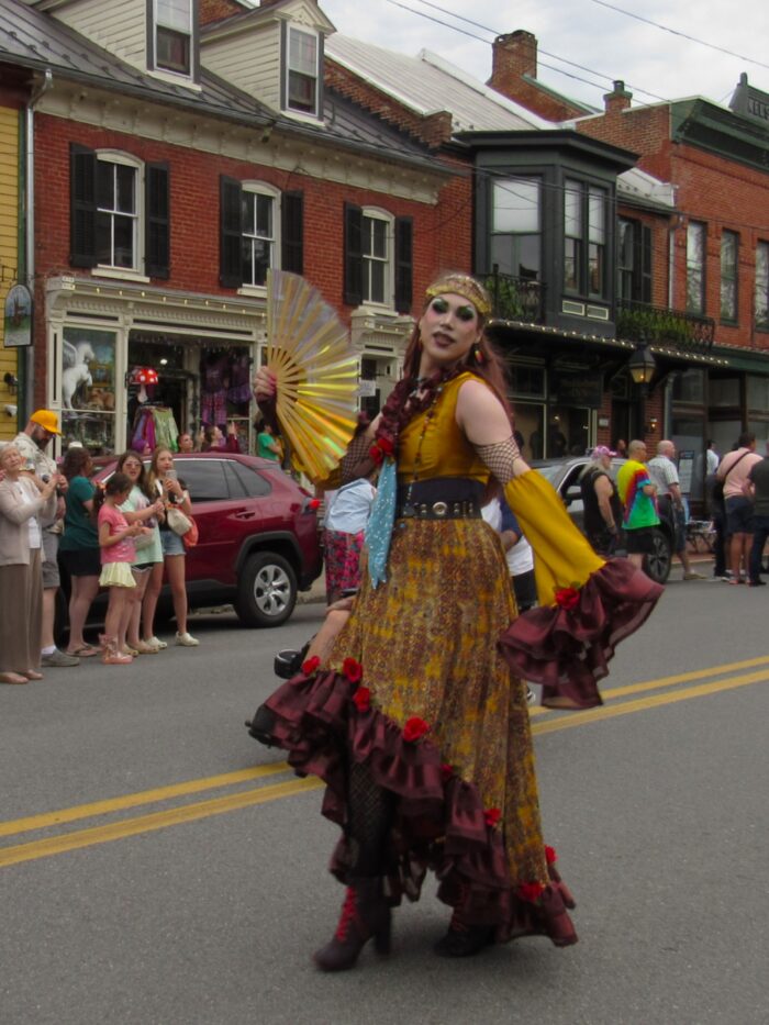 An artist dressed in drag holds a fan and poses for a photograph on a city street as people on the sidewalks look onward.