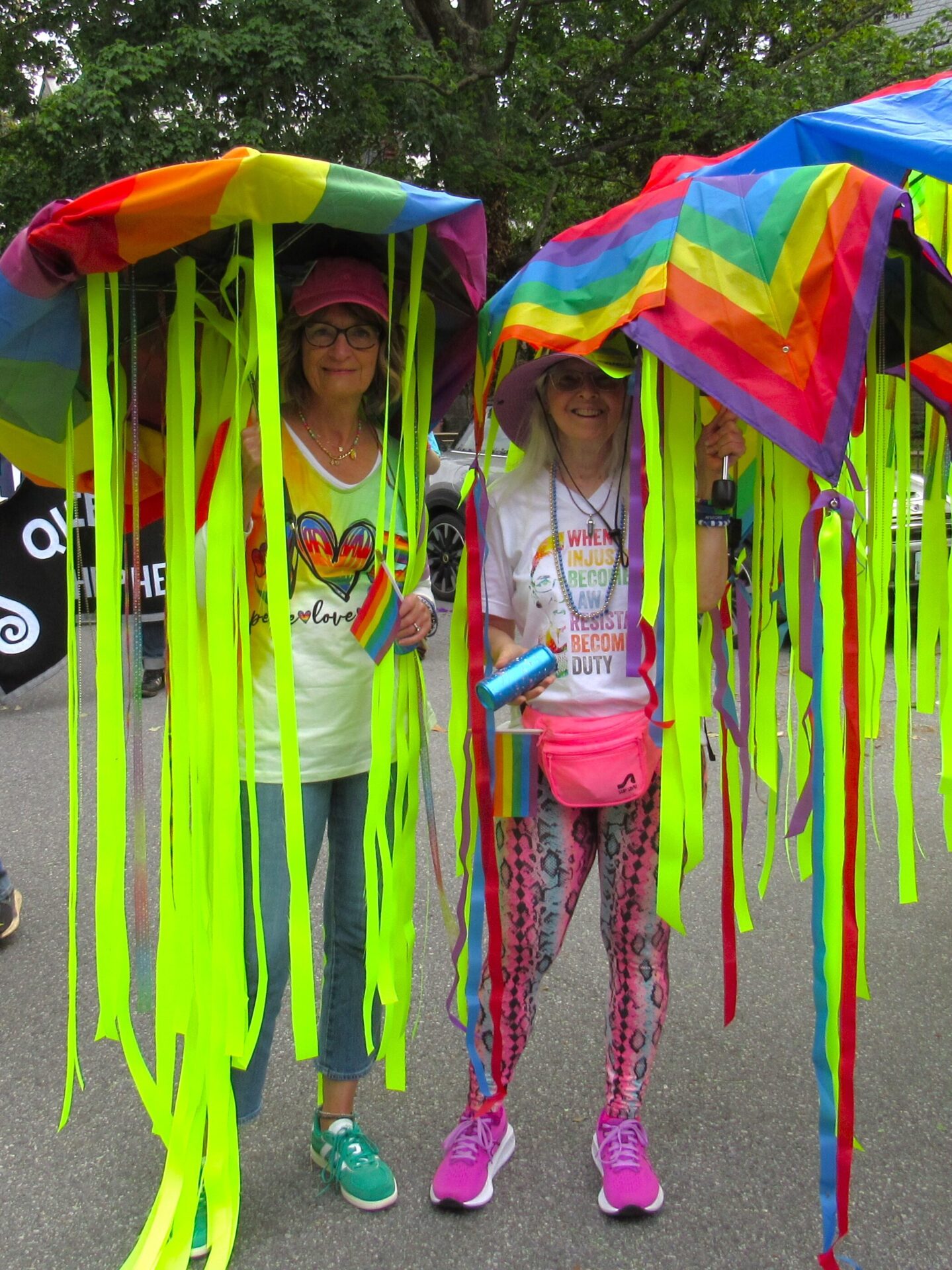 Two people hold up rainbow-colored umbrellas with streamers falling down from the sides, forming a makeshift jellyfish costume.