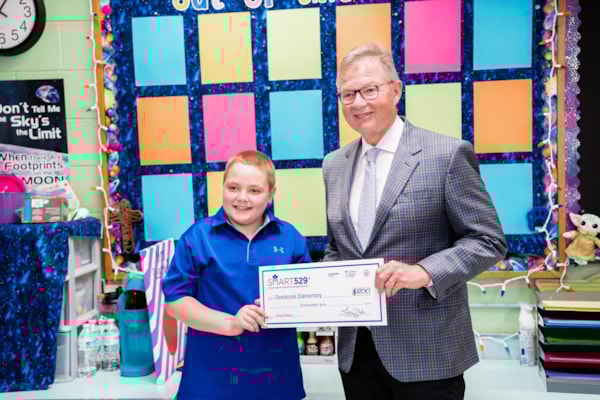 A young boy in a blue polo stands to the left of a man in a grey checkered suit jacket over a white shirt and grey tie. Between them they hold a ceremonial check for $500. The pair stands in front of a classroom cork board decorated with colorful paper.