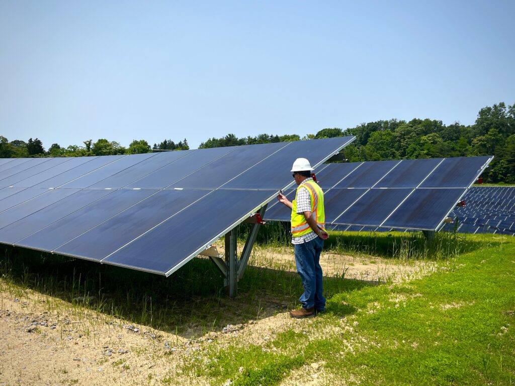 A man in a hard hat and yellow vest stands beside a large solar panel and tweaks something on its outer edge.