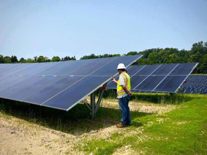 A man in a hard hat and yellow vest stands beside a large solar panel and tweaks something on its outer edge.