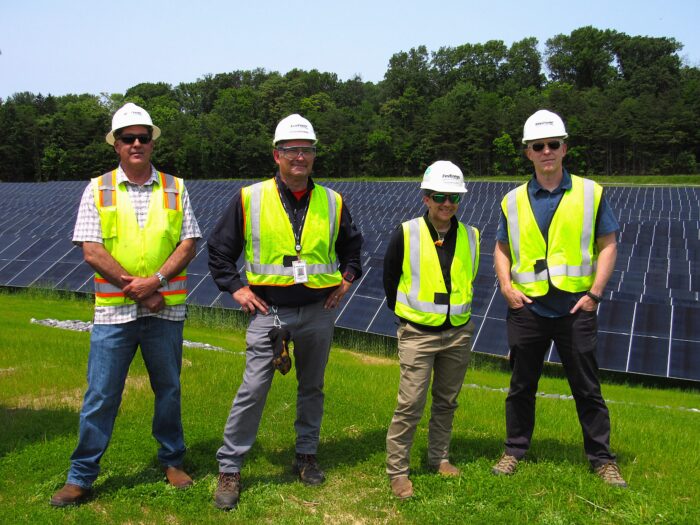 Four people in neon yellow vests and hard hats stand before several rows of solar panels placed on a grassy hill.