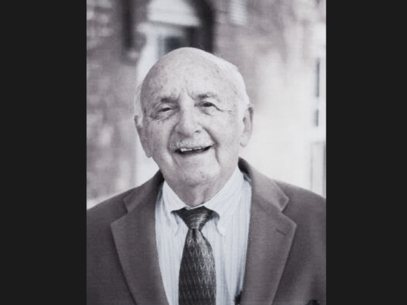 A black-and-white photo shows a man in a suit and tie smiling at the camera for a portrait-style photo. Behind him is a building wall.