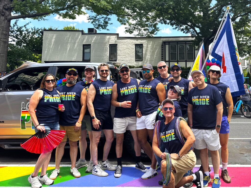 A group of people wearing blue shirts that read "Lost River Pride." They stand on a road painted in rainbow colors, and stand before a pickup truck that displays West Virginia and LGBTQ Pride flags on the back.