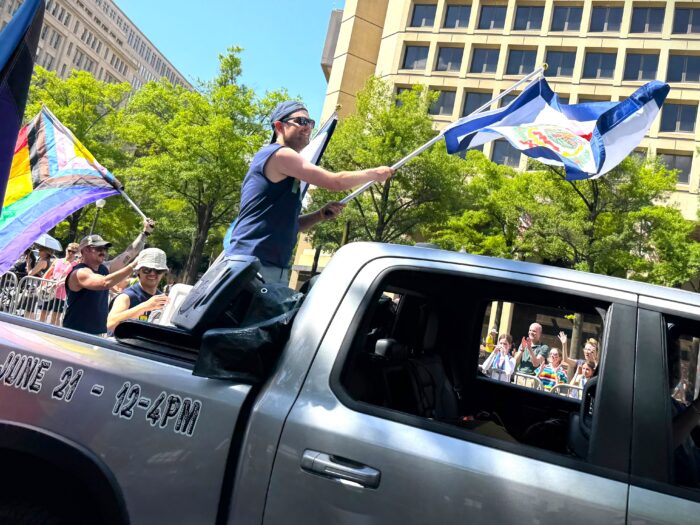 A man stands in the bed of a pickup truck and waves a West Virginia flag. Behind the truck, onlookers line the street and watch on.