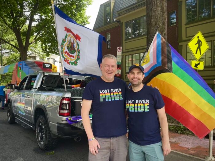 Two men stand outside a pickup truck parked on a city street, which waves West Virginia and LGBTQ Pride flags from its back. The men are smiling at the camera and wear shirts that read "Lost River Pride."