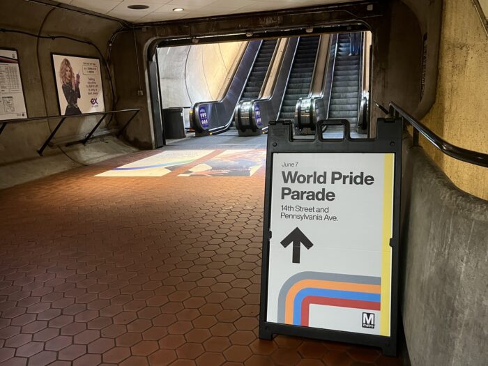 A sign placed before an underground hallway leading to an escalator reads "WorldPride Parade" and displays and arrow pointing straight ahead.