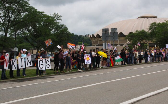 A crowd holds signs in front of the West Virginia University Coliseum as a line on a street. On the far left a group of people hold up individual signs that together spell "NO KINGS". Another person holds a sign that has a red circle and line through a crown. Others wave the flag of the United States under an overcast sky.