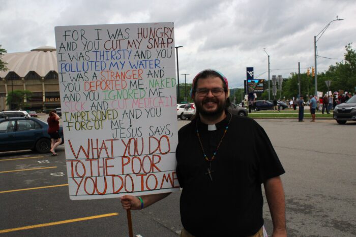 A bearded man wearing a clerical collar on a black shirt, a cross made of nails, a tie dye bandana in his hair and glasses holds a large sign to his left that reads "For I was hungry and you cut my SNAP. I was thirsty and you polluted my water, I was a stranger and you deported me, naked and you ignored me, sick and cut Medicaid, imprisoned and you forgot me! Jesus says, what you do to the poor you do to me." Behind him can be seen the West Virginia University Coliseum