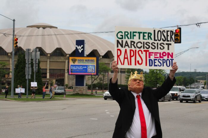 A man wearing a black suit and a red tie over a white button up shirt, as well as a crown over a mask resembling Pres. Donald Trump holds a sign that read ‘Grifter, Loser Narcissist, Rapist, Felon, Despot’ in front of the West Virginia University Coliseum. Cars can be seen waiting at a light behind the man and in front of the building.