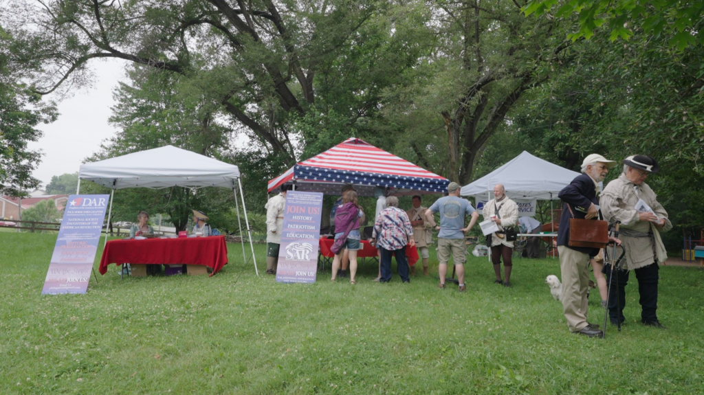 People stand at and around tables beneath tents lined along a grassy park, with signs commemorating the Beeline March.