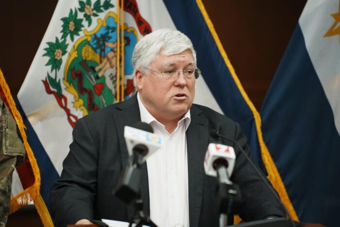 A man with white hair and glasses stands at a podium with microphones. Behind him is the West Virginia flag.