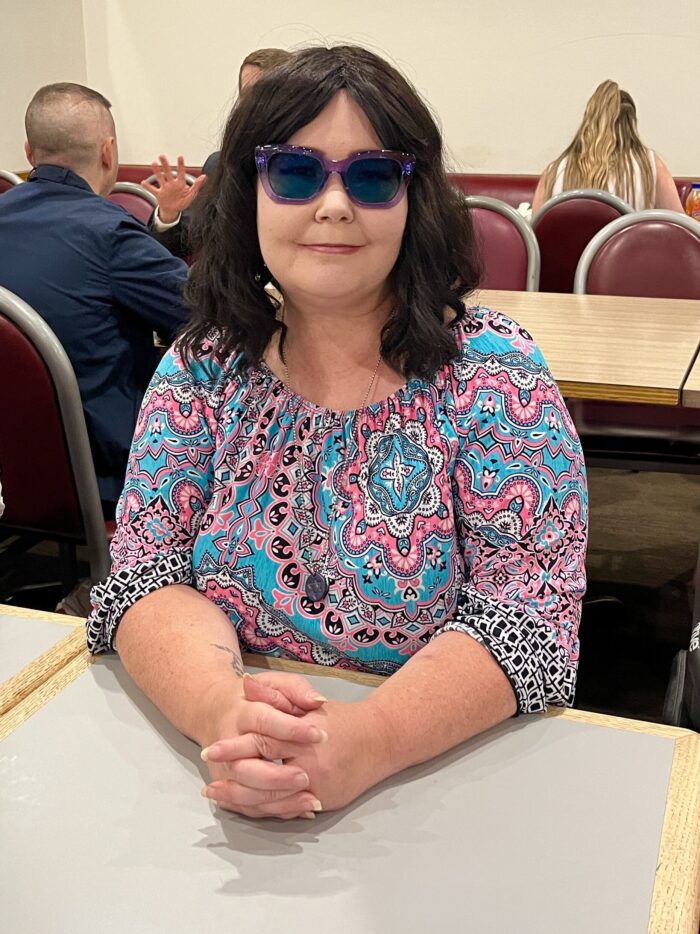 A woman in sunglasses sits at a table and smiles into the camera.