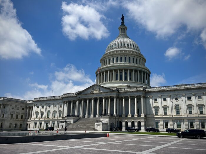 The rotunda and front entryway of the United States Capitol is visible against a partly cloudy sky.